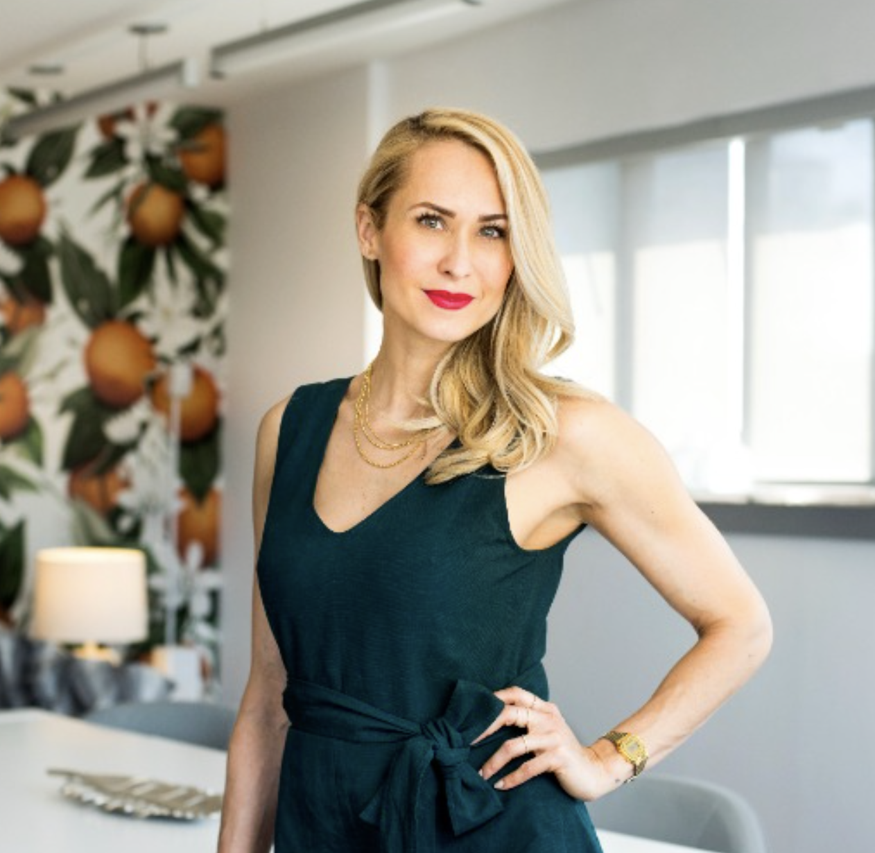 A woman with blonde hair wearing a dark green dress standing with one hand on her hip in a bright room with plants and decor behind her.